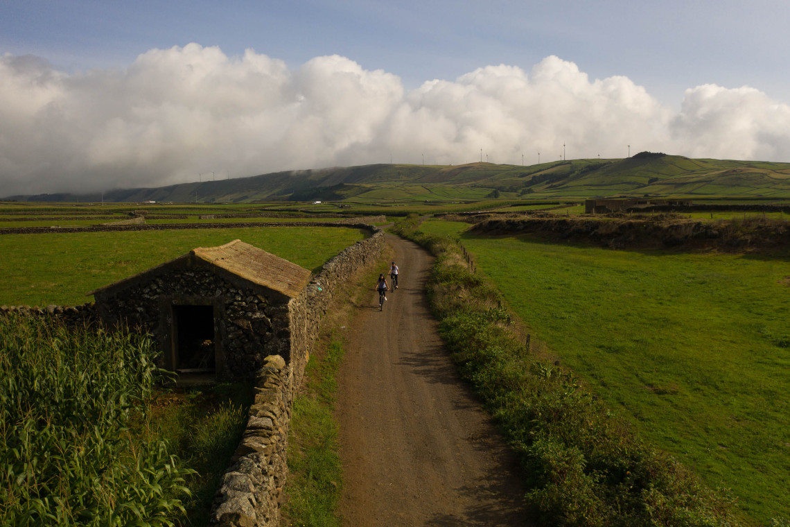 Rural road crossing the green countryside of Terceira Island, surrounded by traditional volcanic stone walls, agricultural fields and a small stone building beneath cloud-covered hills.