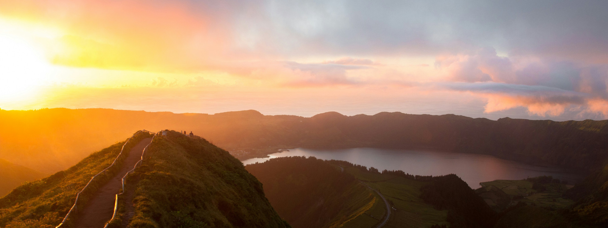 Panoramic view of Lagoa das Sete Cidades at sunset, with dramatic orange and pink sky reflected over the volcanic crater lake and surrounding green hills, São Miguel Island, Azores