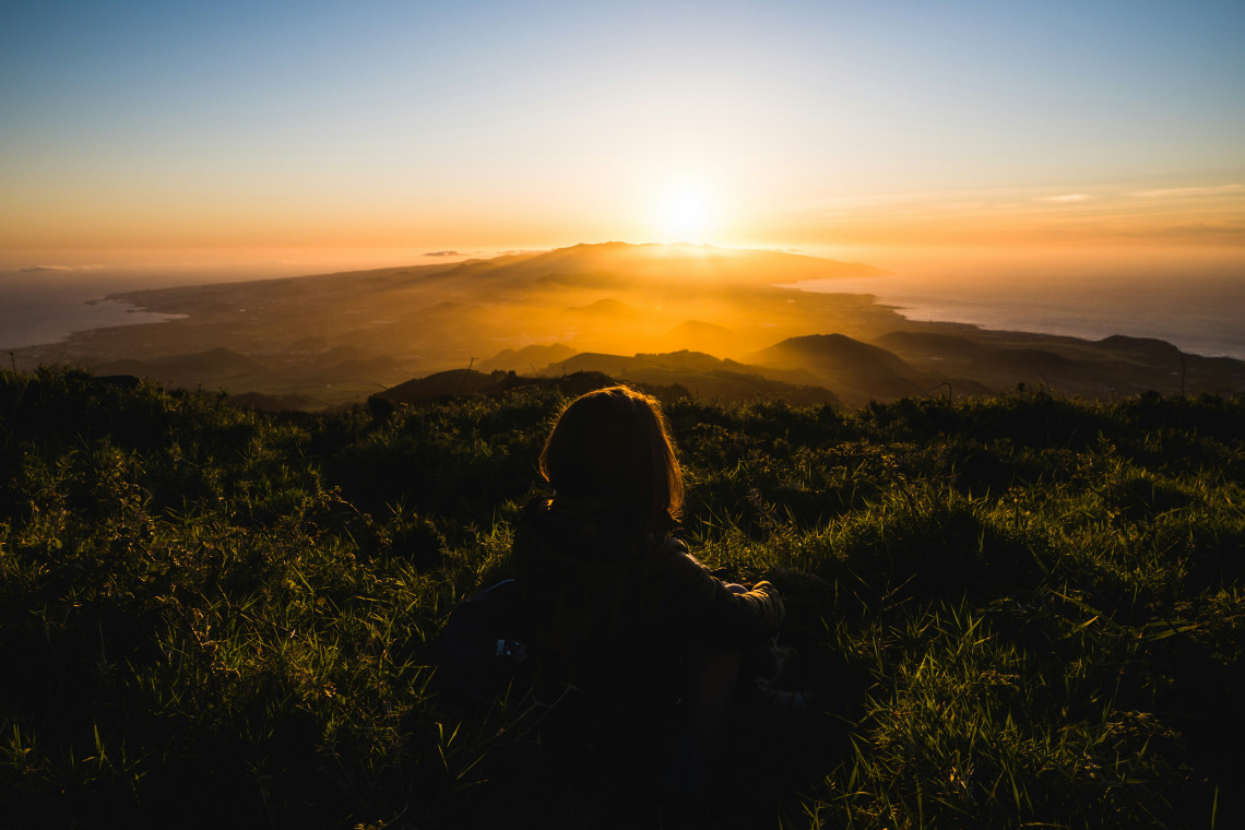 Woman sitting on a hilltop at sunset overlooking Lagoa do Fogo volcanic crater lake, with golden light spreading across São Miguel Island and the Atlantic Ocean in the background, Azores