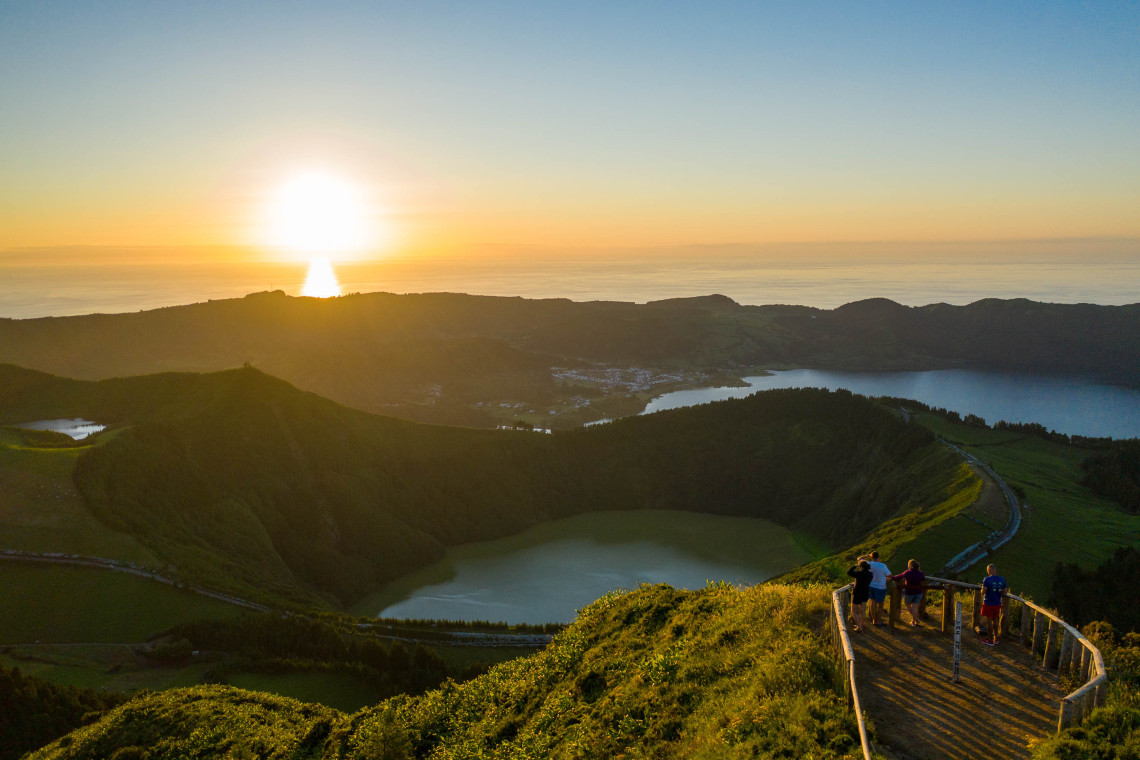 Visitors standing at Miradouro da Grota do Inferno viewpoint looking out over Lagoa das Sete Cidades crater lake at golden hour, with the village and caldera walls visible below, São Miguel Island, Azores