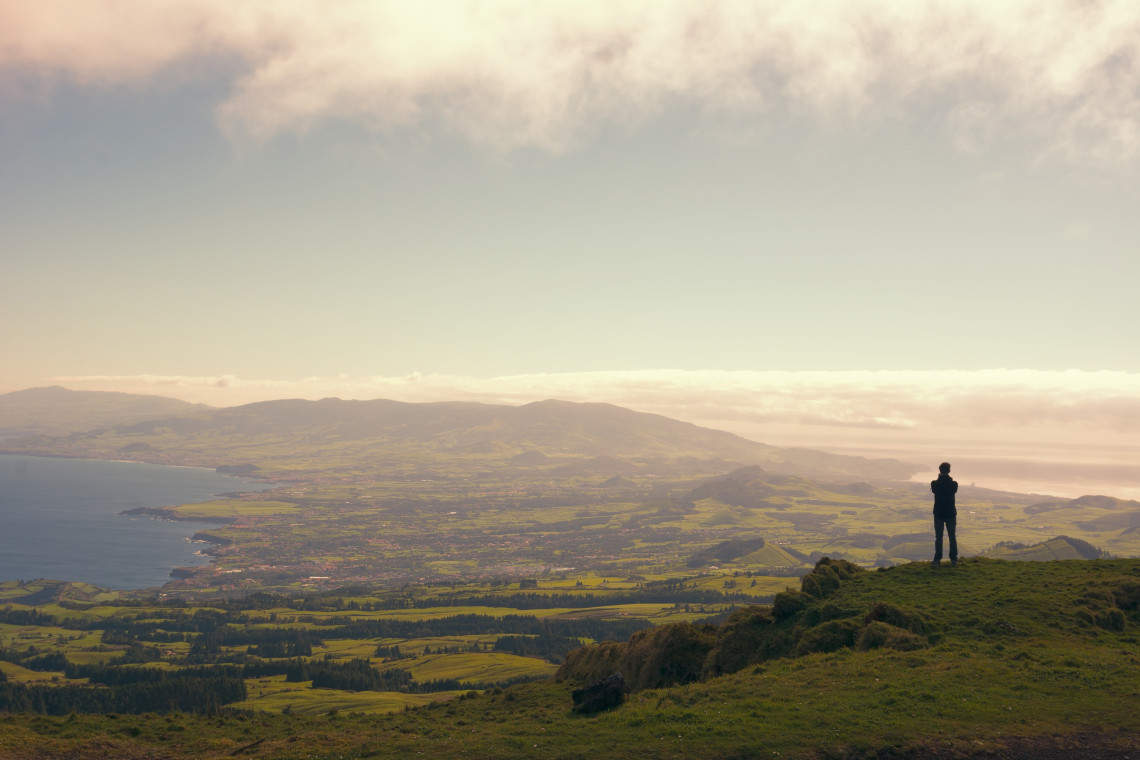Lone traveller standing at Miradouro do Pico do Carvão viewpoint, looking out over the lush green central plateau and Atlantic coastline of São Miguel Island, Azores