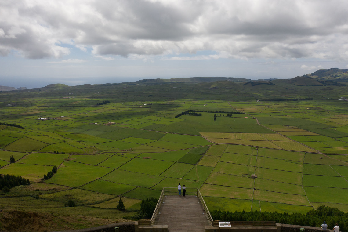 Two visitors standing at Miradouro da Serra do Cume viewpoint, looking out over the iconic mosaic of green fields divided by dark basalt stone walls stretching across Terceira Island, Azores