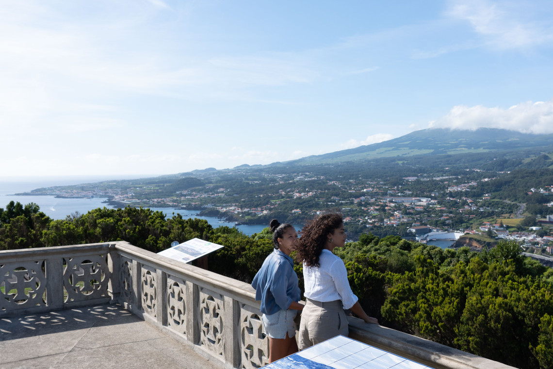 Two visitors at Monte Brasil viewpoint looking out over the rooftops of Angra do Heroísmo, the bay, and Mount Pico rising in the distance across the channel, Terceira Island, Azores