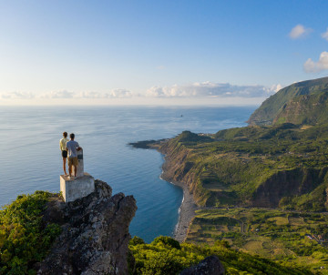 Two visitors standing at Miradouro do Portal viewpoint, looking out over the dramatic green cliffs and Atlantic Ocean coastline of Flores Island, Azores
