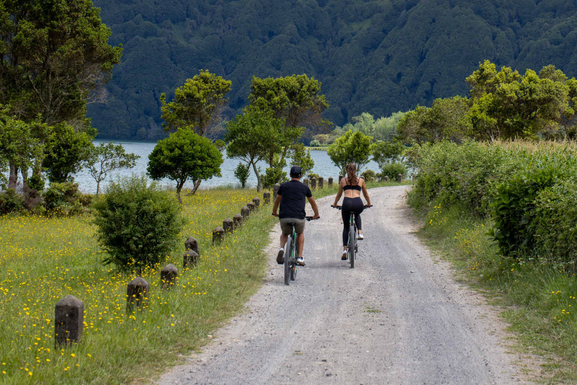 Two cyclists ride along a lakeside path in Sete Cidades on S&atilde;o Miguel Island, surrounded by wildflowers, trees and the steep green walls of the volcanic caldera.