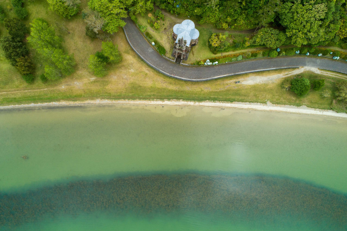 Top-down aerial view of the Chapel of Nossa Senhora das Vit&oacute;rias beside Furnas Lake on S&atilde;o Miguel Island, surrounded by landscaped grounds and forest.
