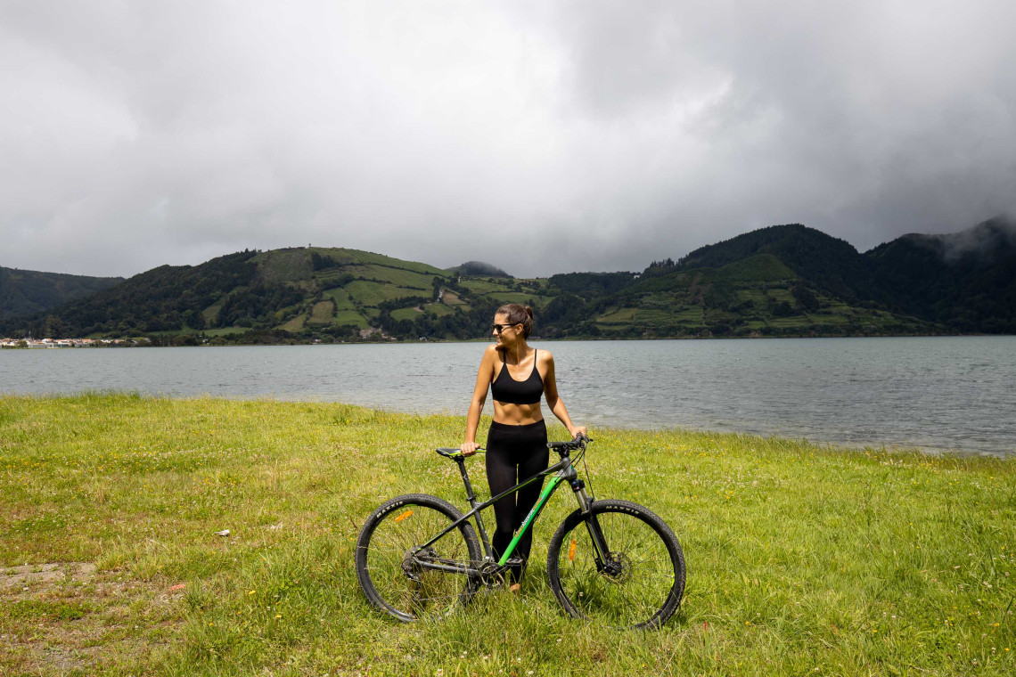 Cyclist standing with a bicycle beside Sete Cidades Lake on S&atilde;o Miguel Island, with green volcanic hills rising beyond the crater lake.