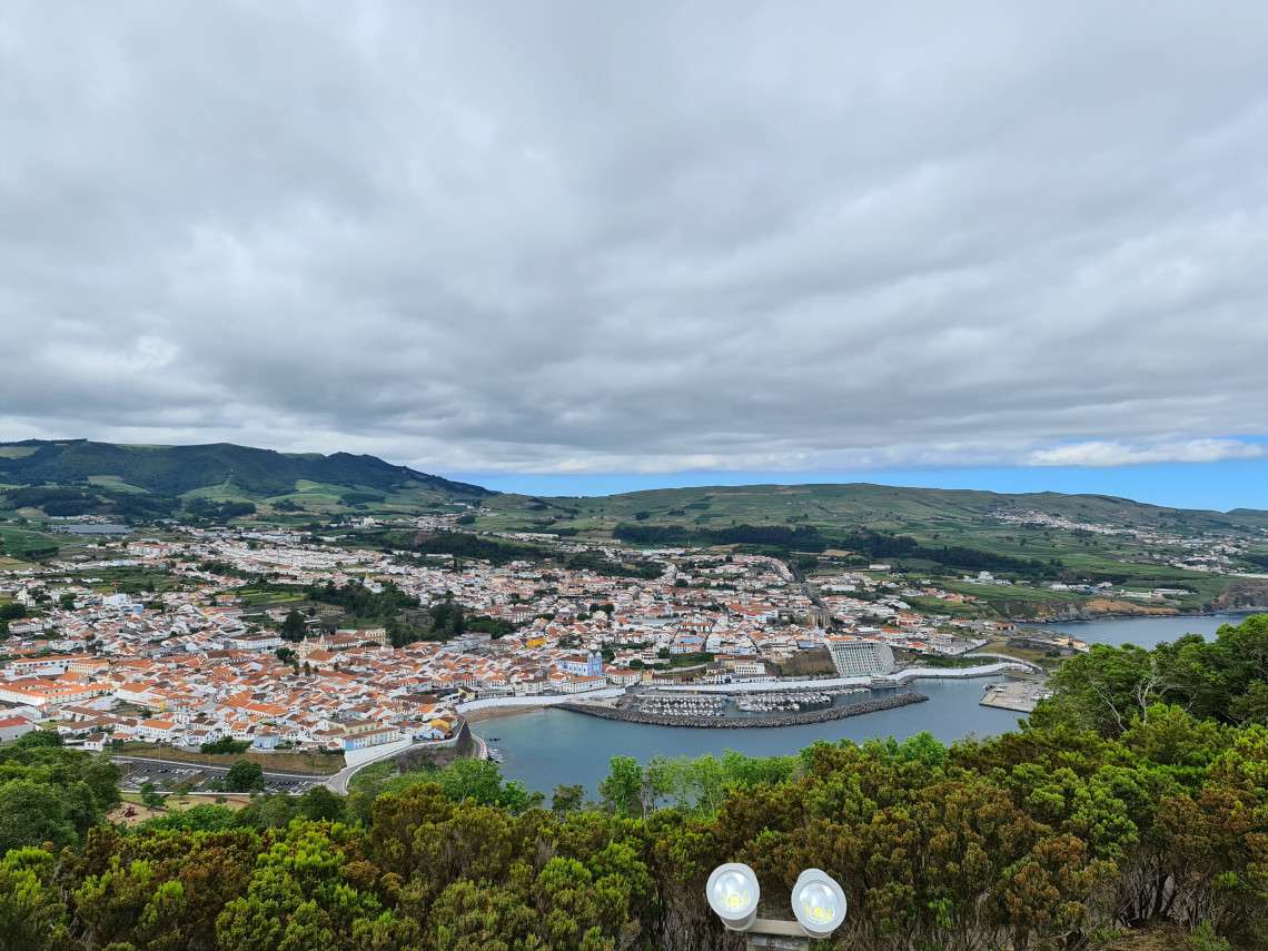 Panoramic view over Angra do Hero&iacute;smo on Terceira Island, showing the red-roofed historic city, harbor and surrounding green hills beneath a cloudy sky.