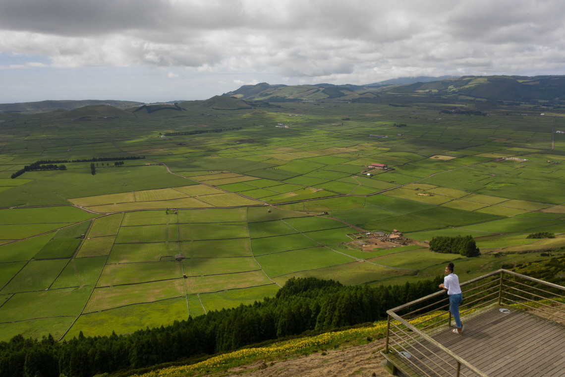 A visitor stands on the Serra do Cume viewpoint overlooking Terceira Island's patchwork of green agricultural fields, rolling hills and distant mountains beneath a cloudy sky.