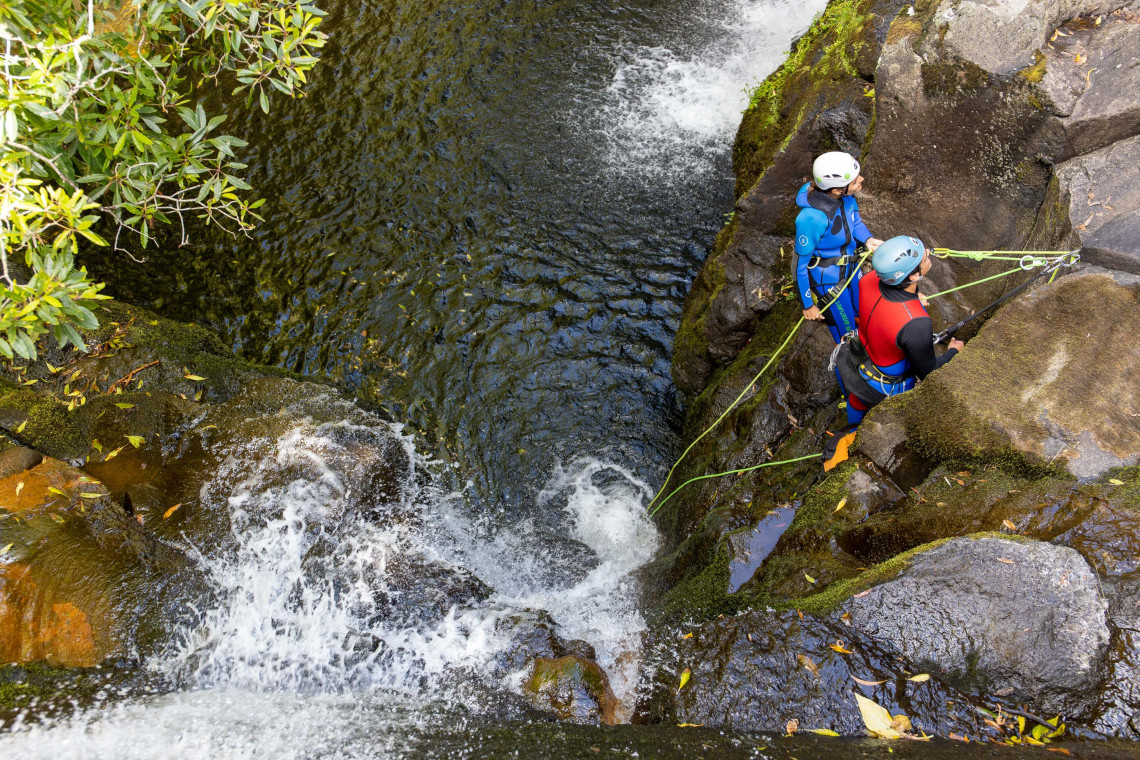 Two canyoning participants prepare to rappel beside a waterfall and dark natural pool on Flores Island in the Azores, Portugal. Ropes are secured to the volcanic rock as part of the guided descent.