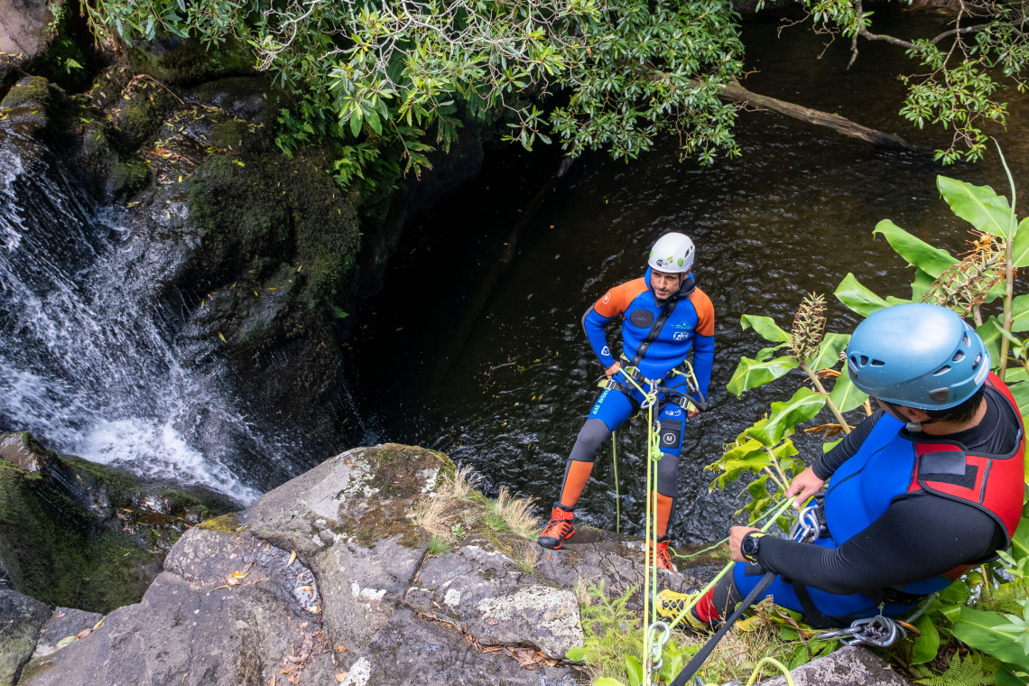 Two canyoning participants use ropes and safety equipment above a waterfall pool on Flores Island in the Azores, Portugal. Moss-covered volcanic rocks and dense vegetation frame the canyon.