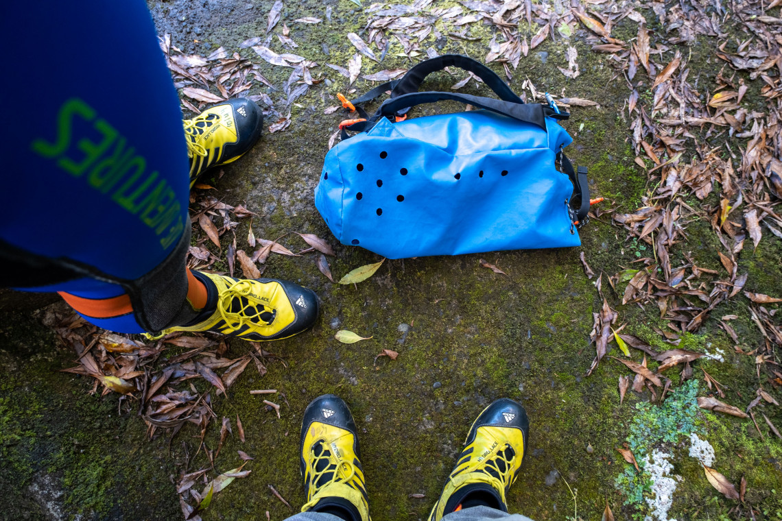 Canyoning boots, wetsuits and a blue waterproof equipment bag rest on the moss-covered forest floor of Flores Island in the Azores, Portugal. The gear is prepared for a guided waterfall and canyon descent.