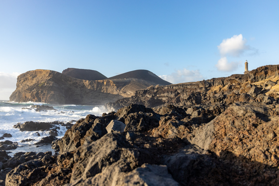 Volcanic rock formations along the Capelinhos coast on Faial Island, Azores, Portugal, with the historic Capelinhos Lighthouse visible above the cliffs.