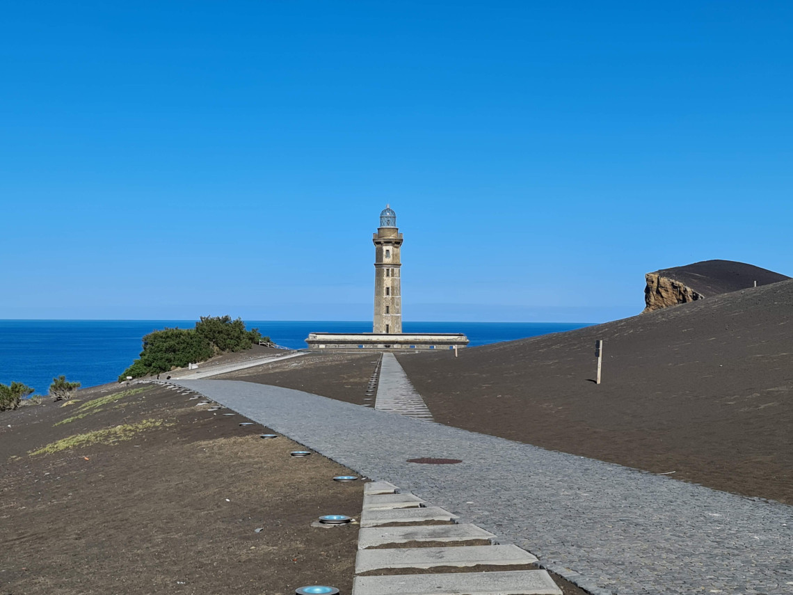 Path leading toward Capelinhos Lighthouse on Faial Island, Azores, Portugal, surrounded by dark volcanic ash with the Atlantic Ocean in the background.