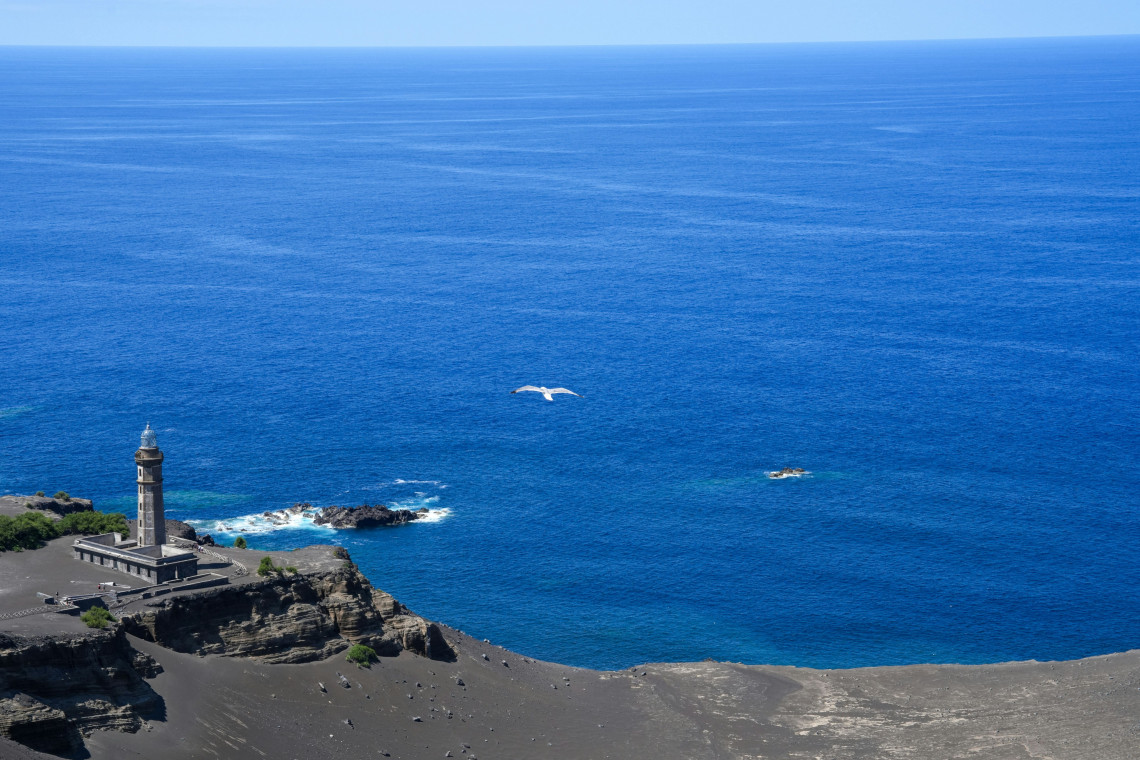 Elevated view of Capelinhos Lighthouse on Faial Island in the Azores, Portugal, surrounded by dark volcanic terrain beside the deep blue Atlantic Ocean.