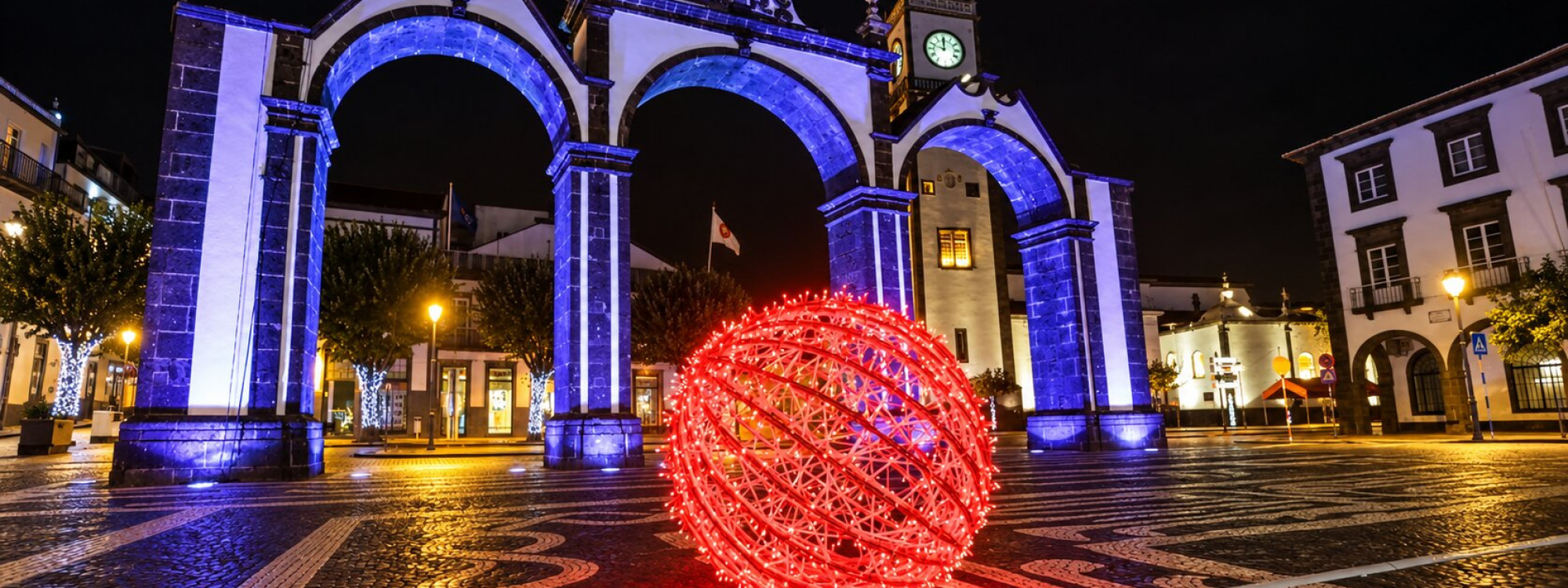 Portas da Cidade illuminated in blue and purple at night in Ponta Delgada, with a large red Christmas ornament displayed in the square.