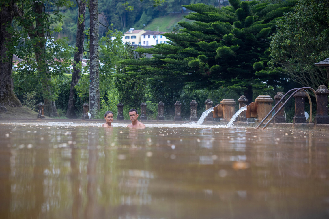 Visitors relax in the warm, iron-rich thermal waters of Terra Nostra Park in Furnas, surrounded by mature trees and historic stone water spouts.