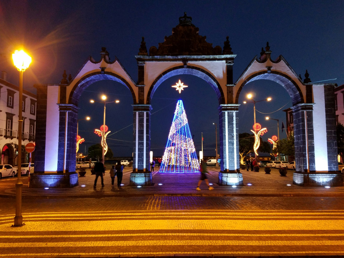 A colorful illuminated Christmas tree stands behind Portas da Cidade in central Ponta Delgada, surrounded by festive street decorations and evening visitors.