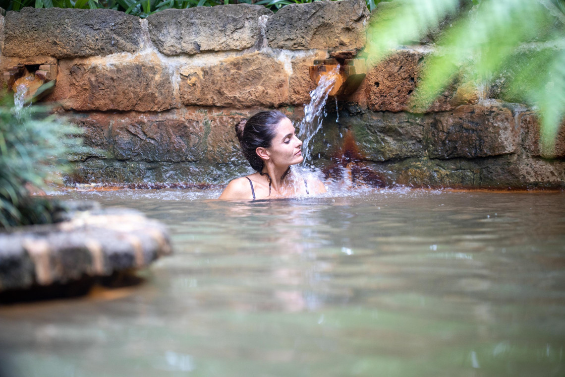 Visitor relaxing in a mineral-rich thermal pool at Terra Nostra Park in Furnas, surrounded by large ferns and dense botanical gardens.