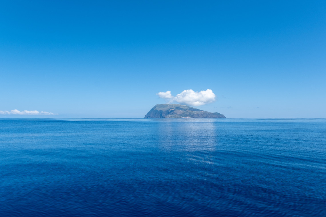 Corvo Island rises from the deep blue Atlantic Ocean beneath a clear sky in the Azores, Portugal. A small cloud rests above the island&rsquo;s volcanic summit while Vila do Corvo appears along the coast.  Image 4