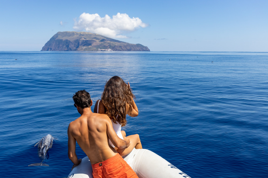 A dolphin surfaces beside a small boat as two travellers observe the marine wildlife near Corvo Island in the Azores, Portugal. The island&rsquo;s volcanic profile extends across the horizon behind them.