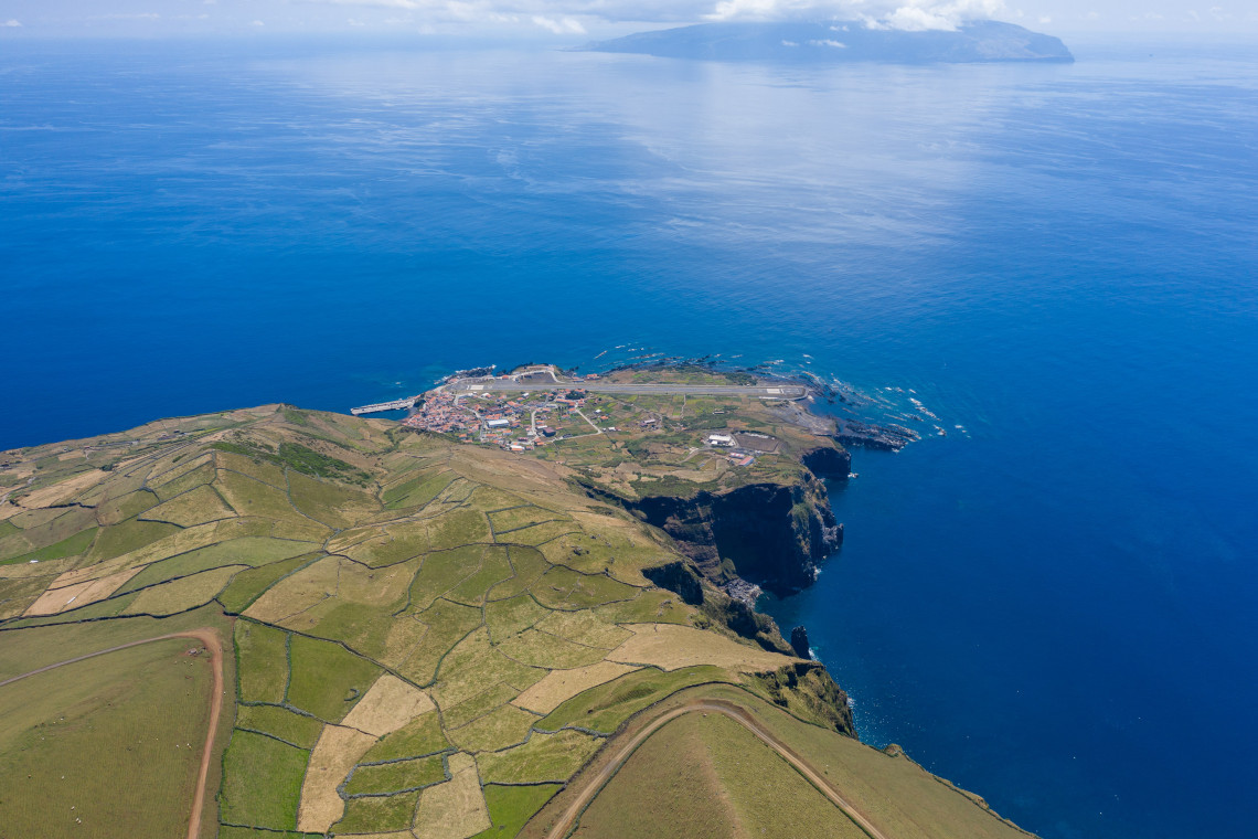 Aerial view of Vila do Corvo and its harbour on the southern coast of Corvo Island in the Azores, Portugal. Patchwork fields and volcanic cliffs extend toward the Atlantic Ocean, with Flores Island visible in the distance.