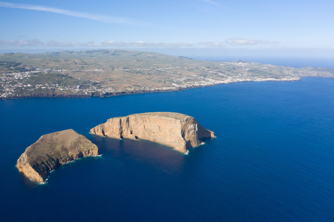 Aerial view of the two volcanic Ilh&eacute;us das Cabras off the southern coast of Terceira Island, surrounded by deep blue Atlantic waters with the island coastline behind them.