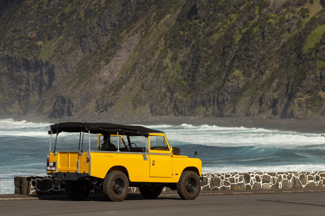 A yellow off-road vehicle is parked beside the Atlantic Ocean, with waves breaking along a dark volcanic beach backed by steep rocky cliffs.