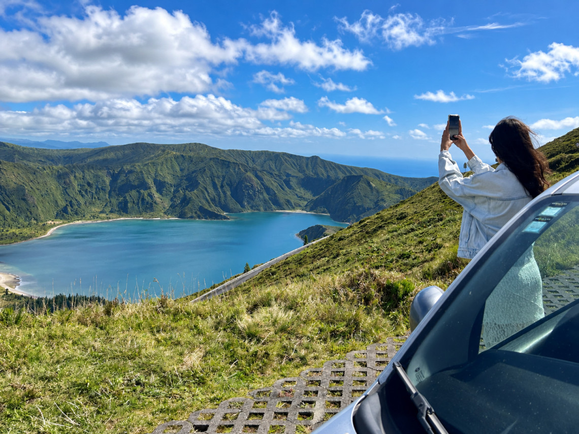 Visitor photographing Lagoa do Fogo from a roadside viewpoint on S&atilde;o Miguel Island, with the blue crater lake and steep green volcanic slopes below.