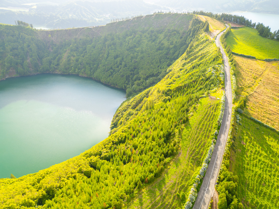 Aerial view of a narrow road crossing a green volcanic ridge in Sete Cidades, with a crater lake on one side and the surrounding caldera landscape beyond.