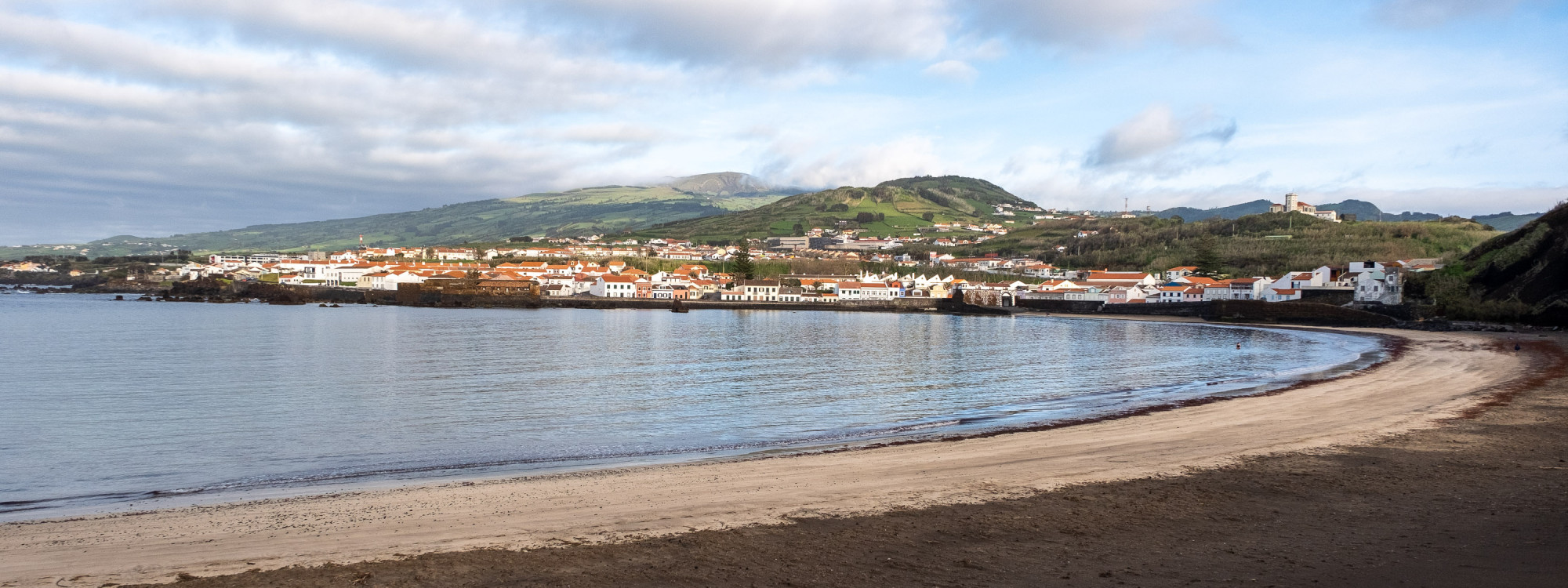 Porto Pim Beach in Horta, Faial, with a curved sandy shoreline, calm Atlantic waters and green hills surrounding the bay.
