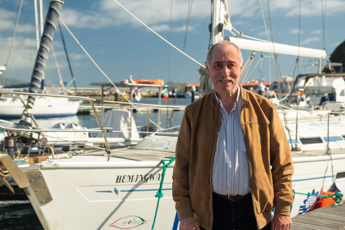 Man standing beside a sailing boat at Horta Marina on Faial Island in the Azores, Portugal, with other yachts and marina infrastructure in the background.