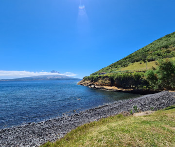 Baía das Canas on Pico Island, with a dark volcanic pebble shoreline, calm Atlantic water and green slopes descending toward the coast.
