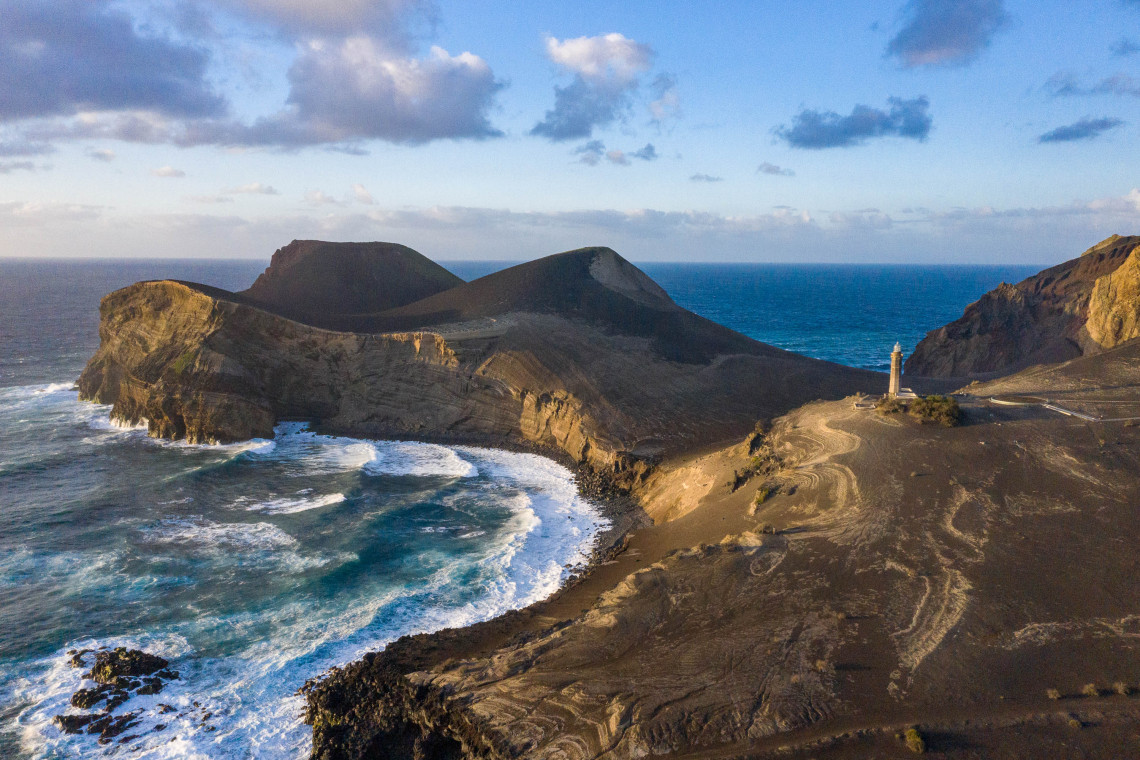 Aerial view of the Capelinhos volcanic landscape on western Faial Island, with dark ash fields, rugged Atlantic cliffs and the Capelinhos Lighthouse.