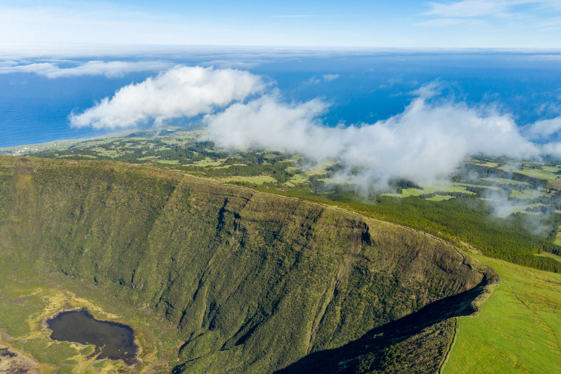 Aerial view across the steep green walls of the Caldeira do Faial, with clouds drifting over the volcanic highlands and the Atlantic Ocean beyond.