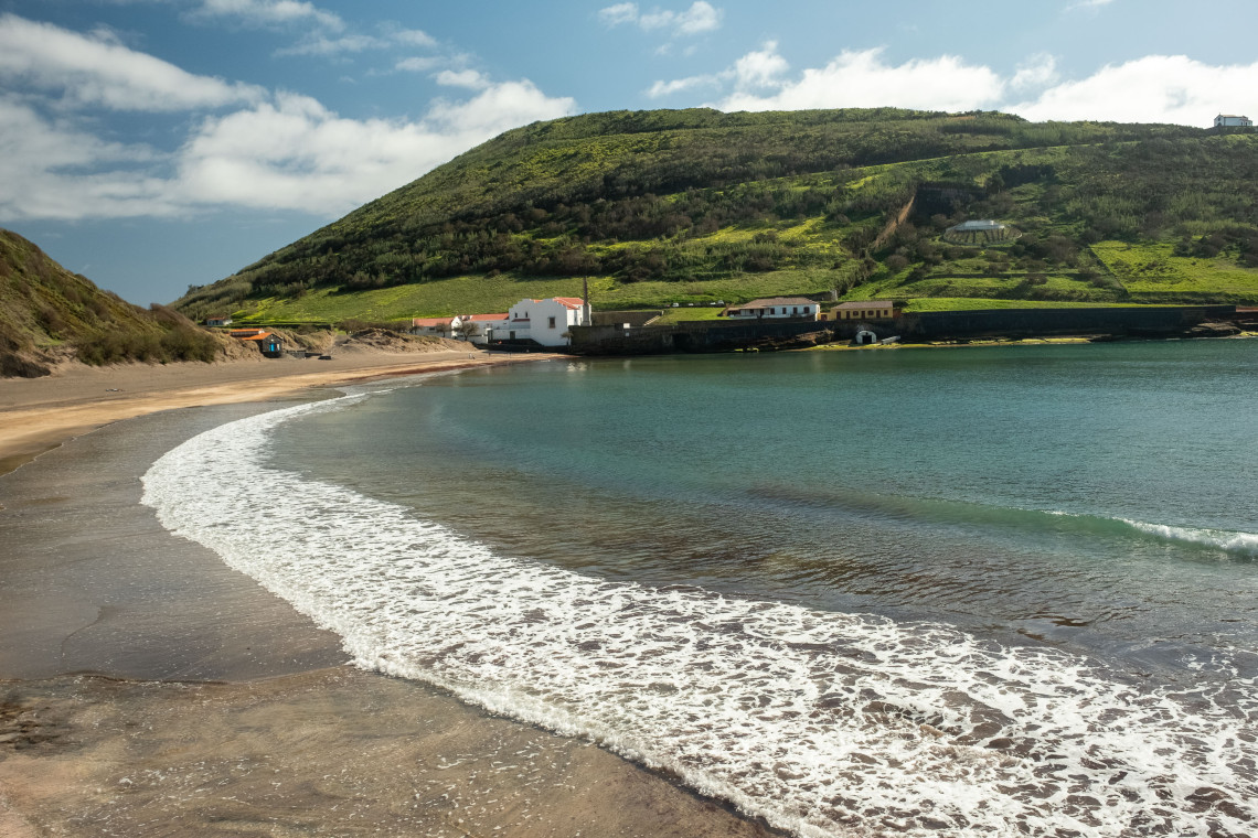 Porto Pim Beach in Horta, with dark volcanic sand, calm Atlantic water and green slopes surrounding the sheltered bay on Faial Island.