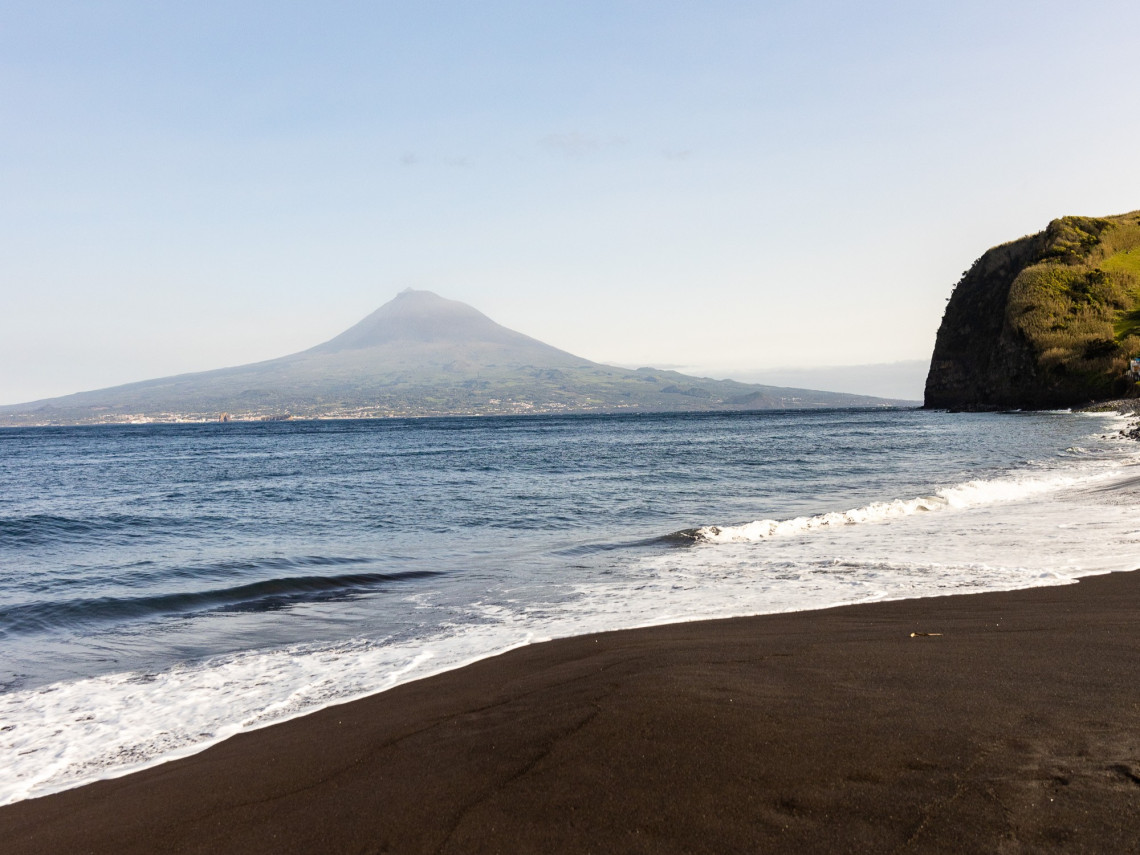 Black volcanic sand at Praia do Almoxarife on Faial Island, with Atlantic waves along the shore and Mount Pico rising across the channel.