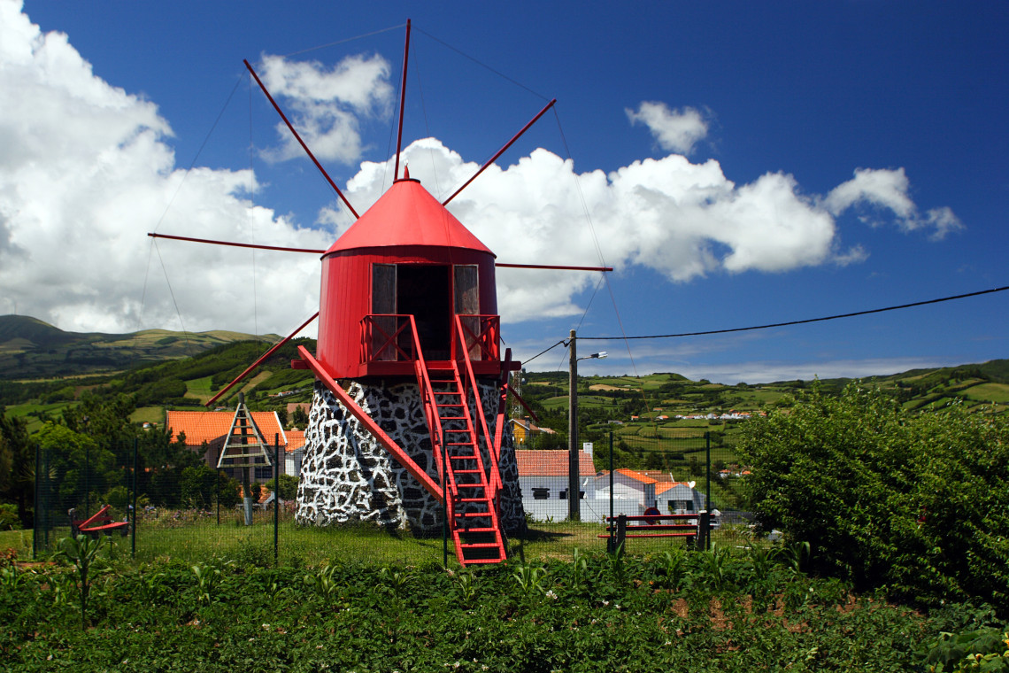 Traditional red windmill at Lomba da Concei&ccedil;&atilde;o in Horta, standing on a volcanic stone base against the green hills and blue sky of Faial Island.