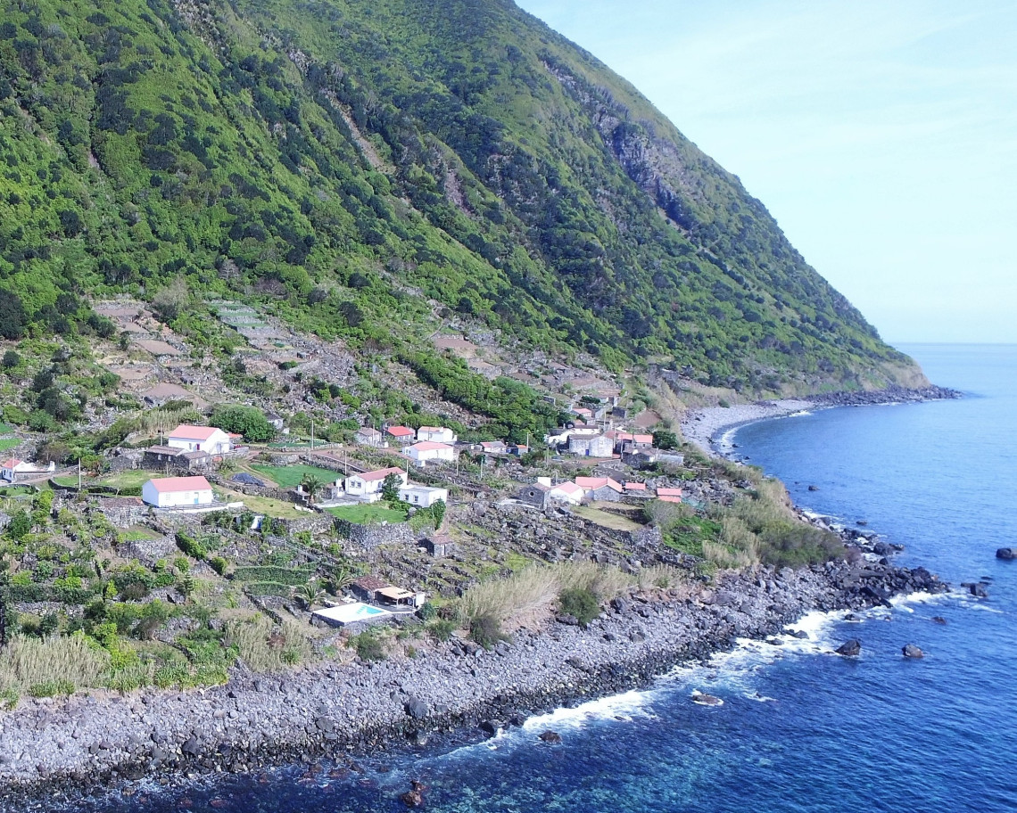 Elevated view of Faj&atilde; dos Bodes on S&atilde;o Jorge Island, with small houses and agricultural plots set between a steep green hillside and the rocky Atlantic coast.