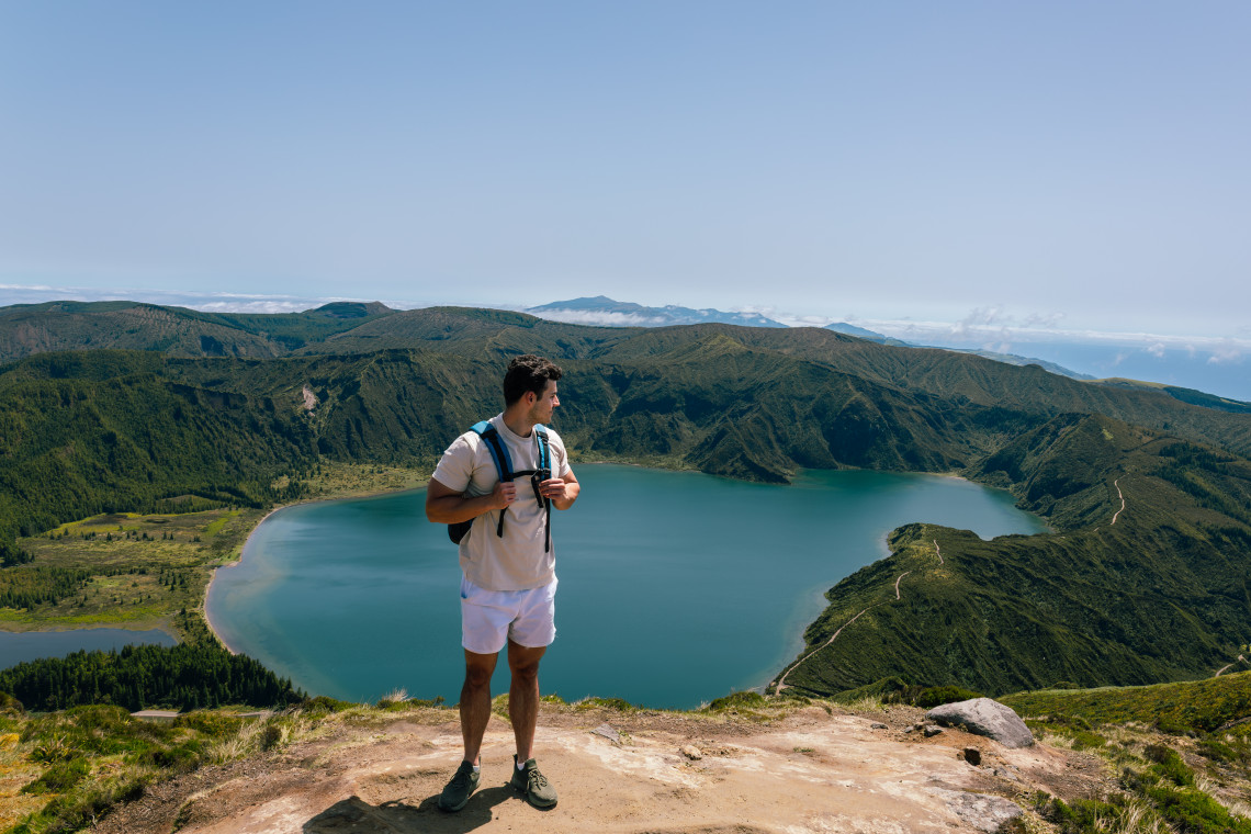 A hiker stands at a viewpoint overlooking Lagoa do Fogo, a volcanic crater lake surrounded by steep green ridges and mountainous terrain on S&atilde;o Miguel Island.