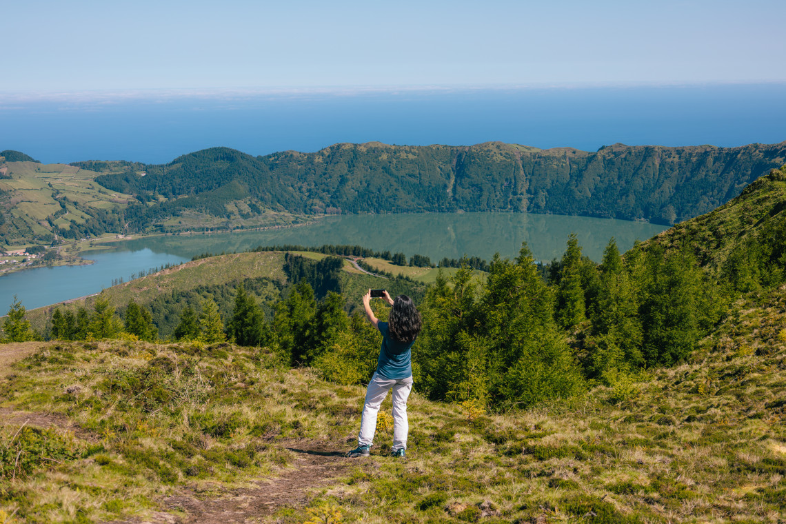 A visitor photographs Lagoa do Fogo from a high viewpoint on S&atilde;o Miguel Island, with forested slopes, volcanic crater walls and the Atlantic Ocean visible beyond the landscape.