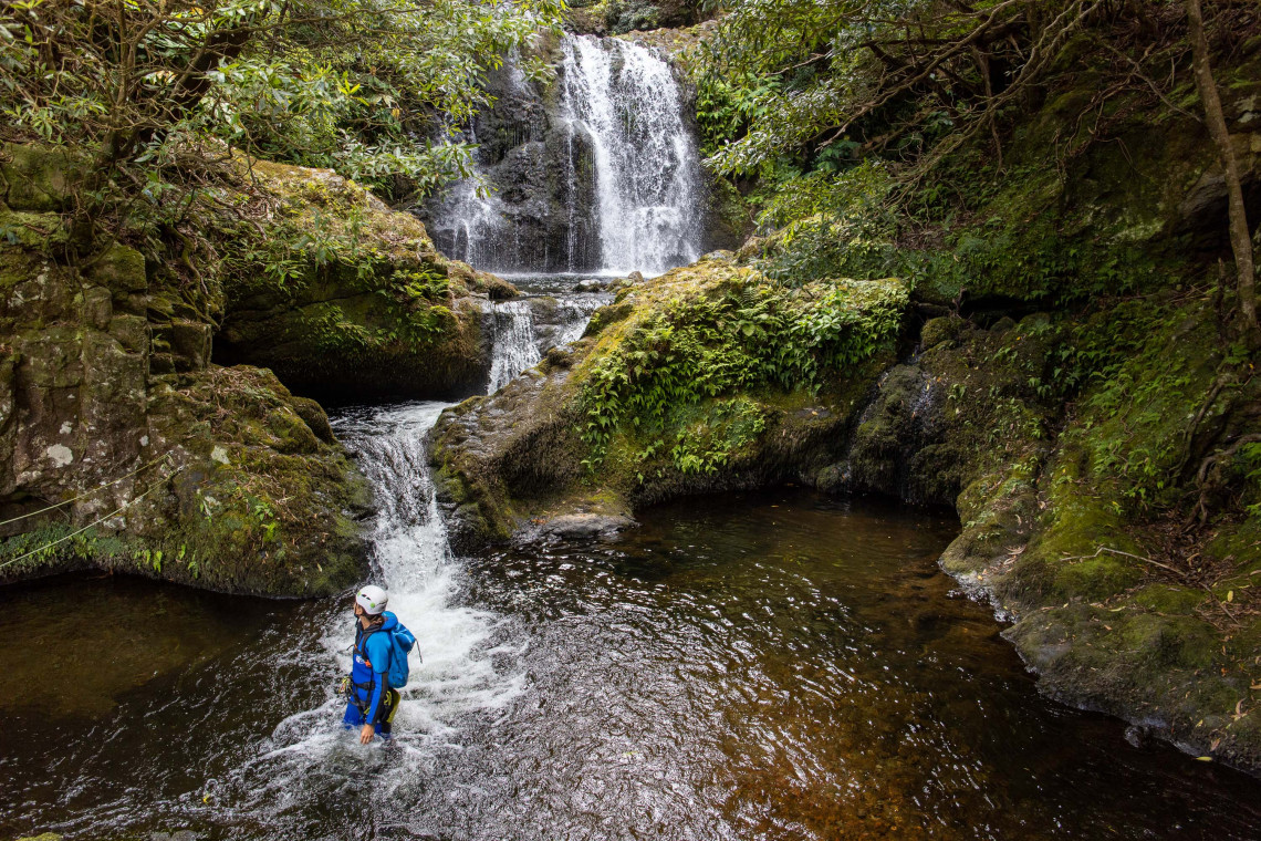 A canyoning participant moves through a cascading stream at Ribeira da Fazenda in Santa Cruz das Flores, Portugal. Moss-covered volcanic rocks and dense forest vegetation surround the waterfall route on Flores Island.