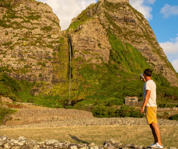 A visitor looks toward the Poço do Bacalhau waterfall in Fajã Grande on Flores Island, Azores. The narrow cascade descends between steep volcanic cliffs covered with dense green vegetation.