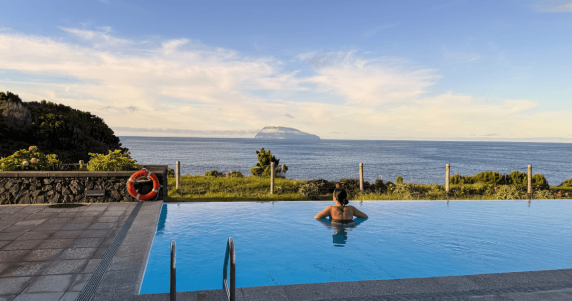 A guest relaxes in the outdoor pool at Ocean Cliff Hotel in Santa Cruz das Flores, Azores, Portugal. The pool overlooks the Atlantic Ocean, with Corvo Island visible on the horizon.