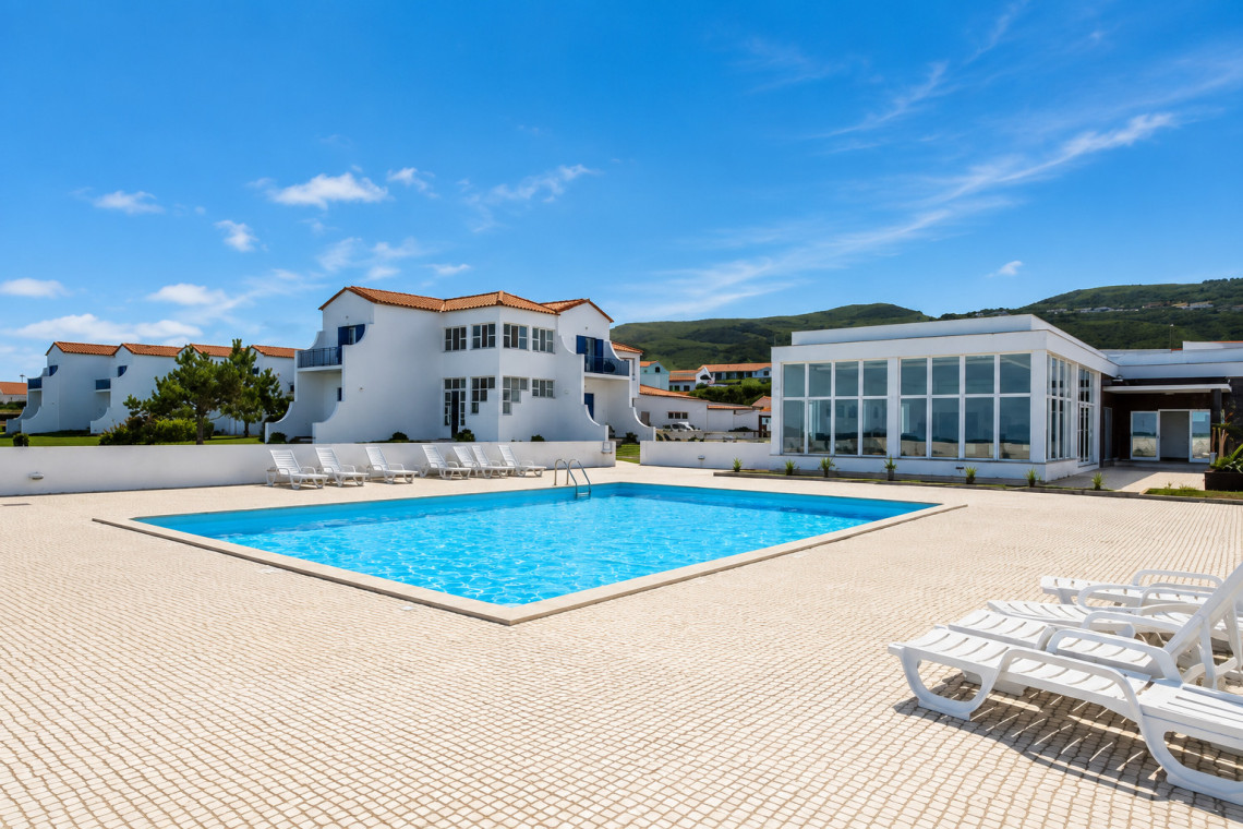 Outdoor swimming pool at Hotel Ocidental in Santa Cruz das Flores on Flores Island, Azores, Portugal. White hotel buildings and sun loungers surround the pool beneath a clear blue sky.