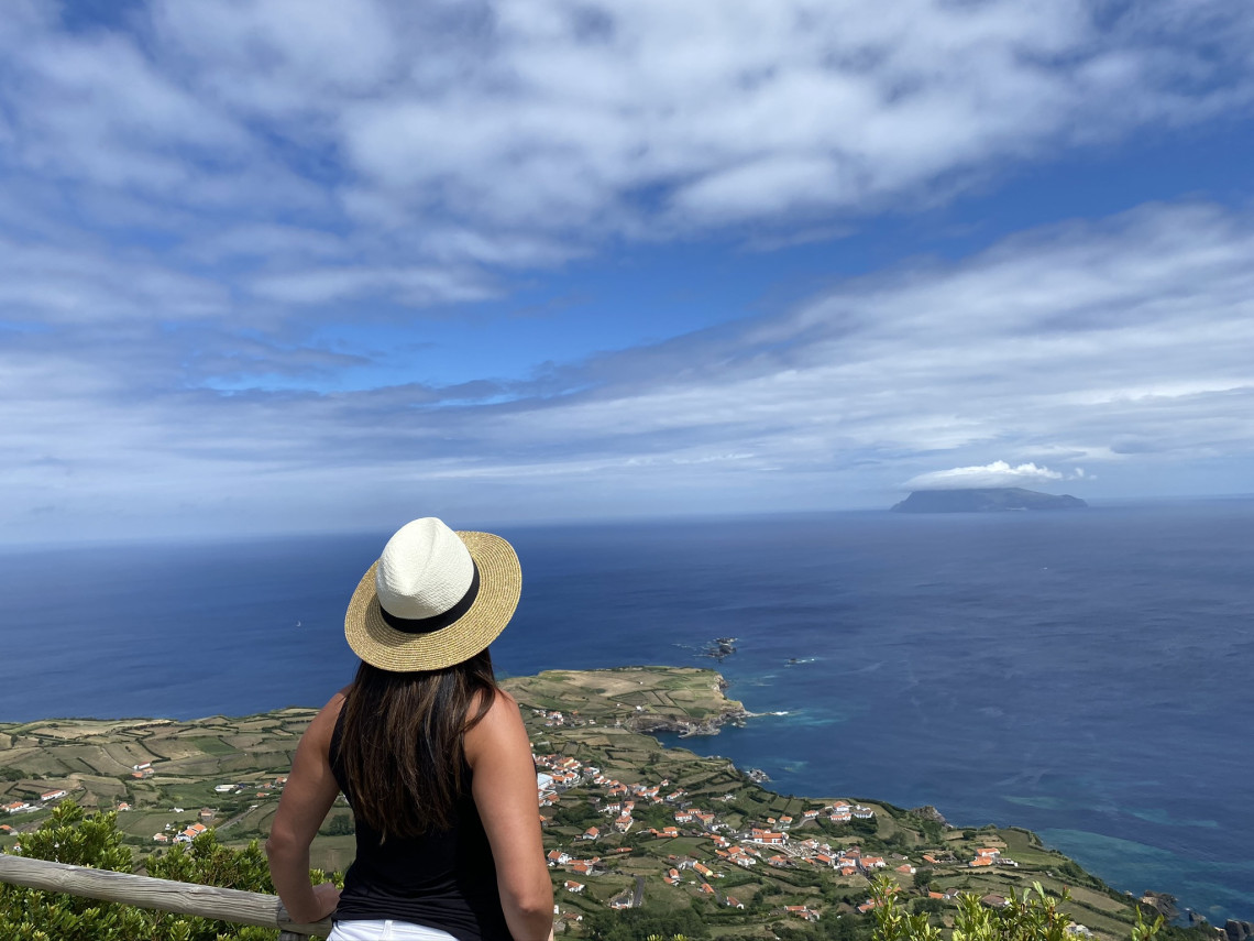 A visitor overlooks the village of Ponta Delgada and its patchwork coastal fields from a viewpoint on Flores Island, Azores, Portugal. Corvo Island rises from the Atlantic Ocean beneath a partly cloudy sky.