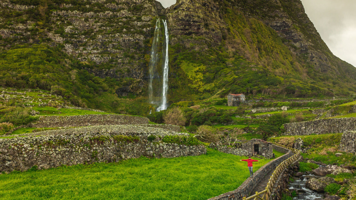 Po&ccedil;o do Bacalhau waterfall descends from a steep volcanic cliff above Faj&atilde; Grande on Flores Island, Portugal. Traditional stone walls, green fields and a small stream surround the base of the waterfall.