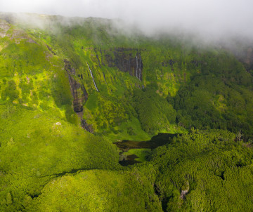 Aerial view of Poço da Ribeira do Ferreiro near Fajã Grande on Flores Island in the Azores, Portugal. Waterfalls descend through steep green volcanic cliffs partially covered by low clouds.