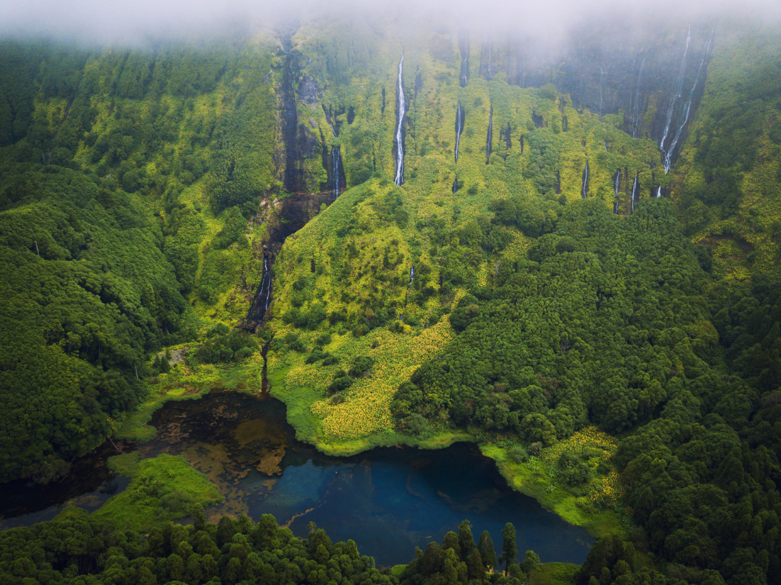 Aerial view of Po&ccedil;o da Ribeira do Ferreiro near Faj&atilde;zinha on Flores Island, Azores, Portugal. Several waterfalls descend through a mist-covered green cliff toward a dark lagoon surrounded by dense forest.