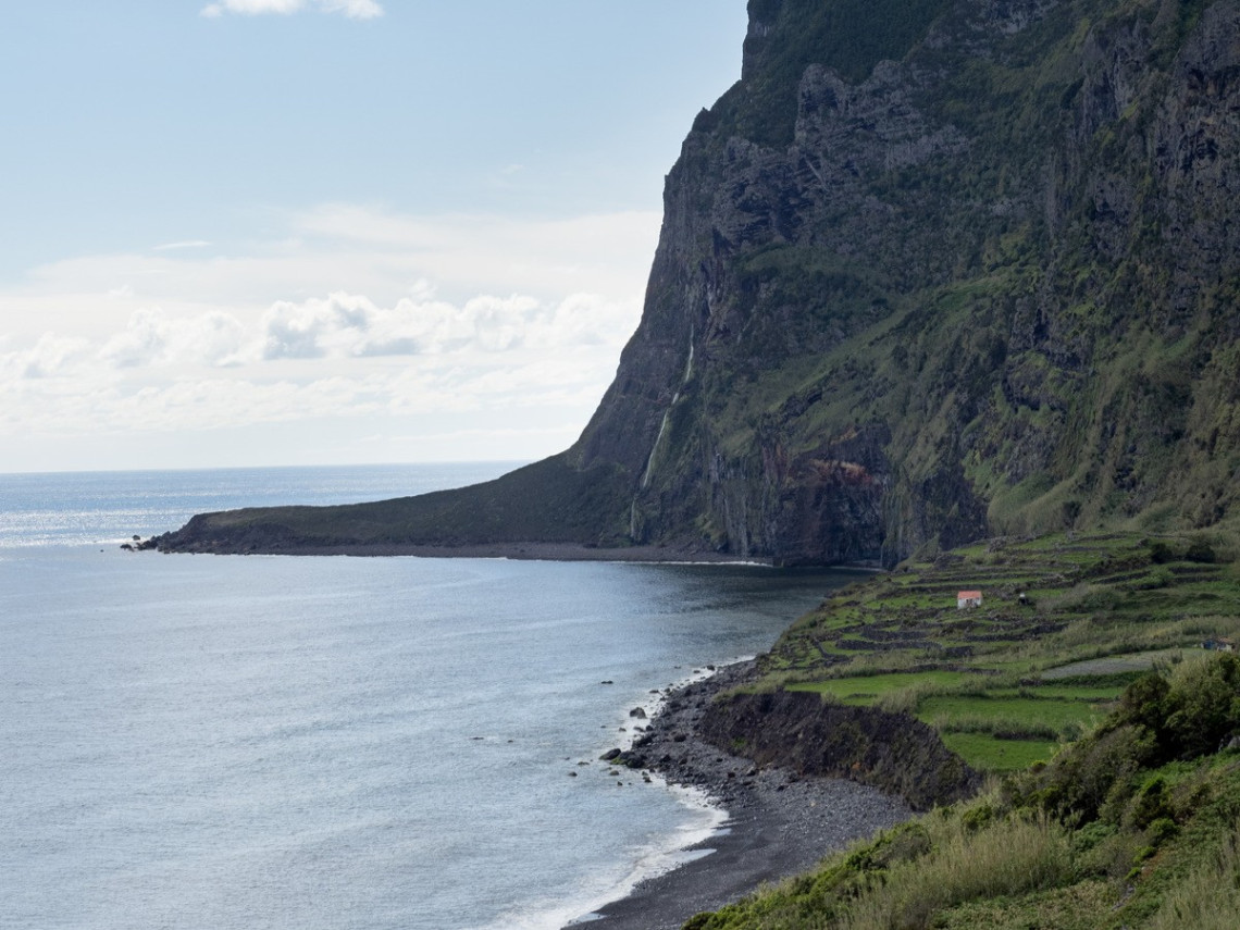 Faj&atilde; de Lopo Vaz on the southern coast of Flores Island, Azores, Portugal, with terraced farmland beneath a steep volcanic cliff. A dark pebble beach curves around the sheltered Atlantic bay.